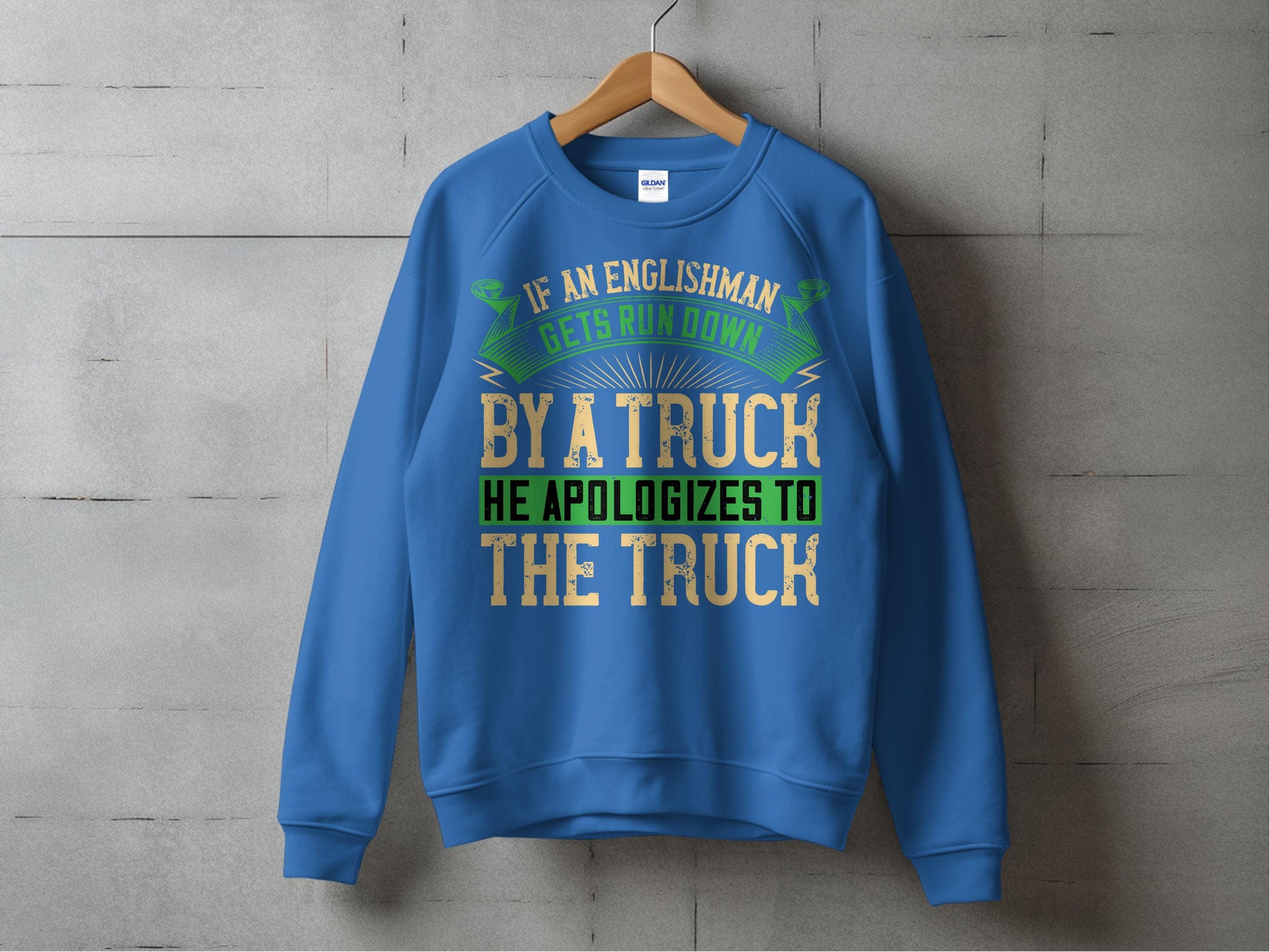A blue sweatshirt from Garment Graphics on a wooden hanger against a concrete wall features the humorous text, If an Englishman gets run down by a truck, he apologizes to the truck, in yellow, green, and white fonts. Its available in unisex sizes for versatile wear.