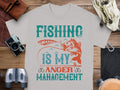 The Garment Graphics Fishing is my anger management T-shirt for fishing enthusiasts, featuring a fish and hook image, lies on a wooden surface surrounded by items like a camera, binoculars, leather accessories, and a coiled rope.