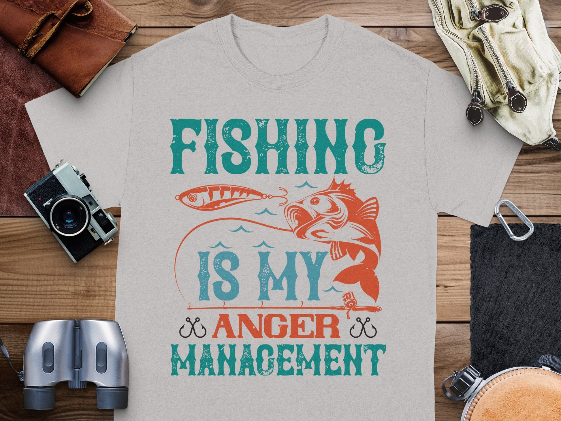 The Garment Graphics Fishing is my anger management T-shirt for fishing enthusiasts, featuring a fish and hook image, lies on a wooden surface surrounded by items like a camera, binoculars, leather accessories, and a coiled rope.