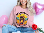 A person holds a red rose while wearing a Garment Graphics pink Bee Yourself, Everyone Else is Taken Summer Edition sweatshirt. Surrounded by three pink heart-shaped balloons and paired with ripped blue jeans, they enjoy the comfy fit of unisex sizing.