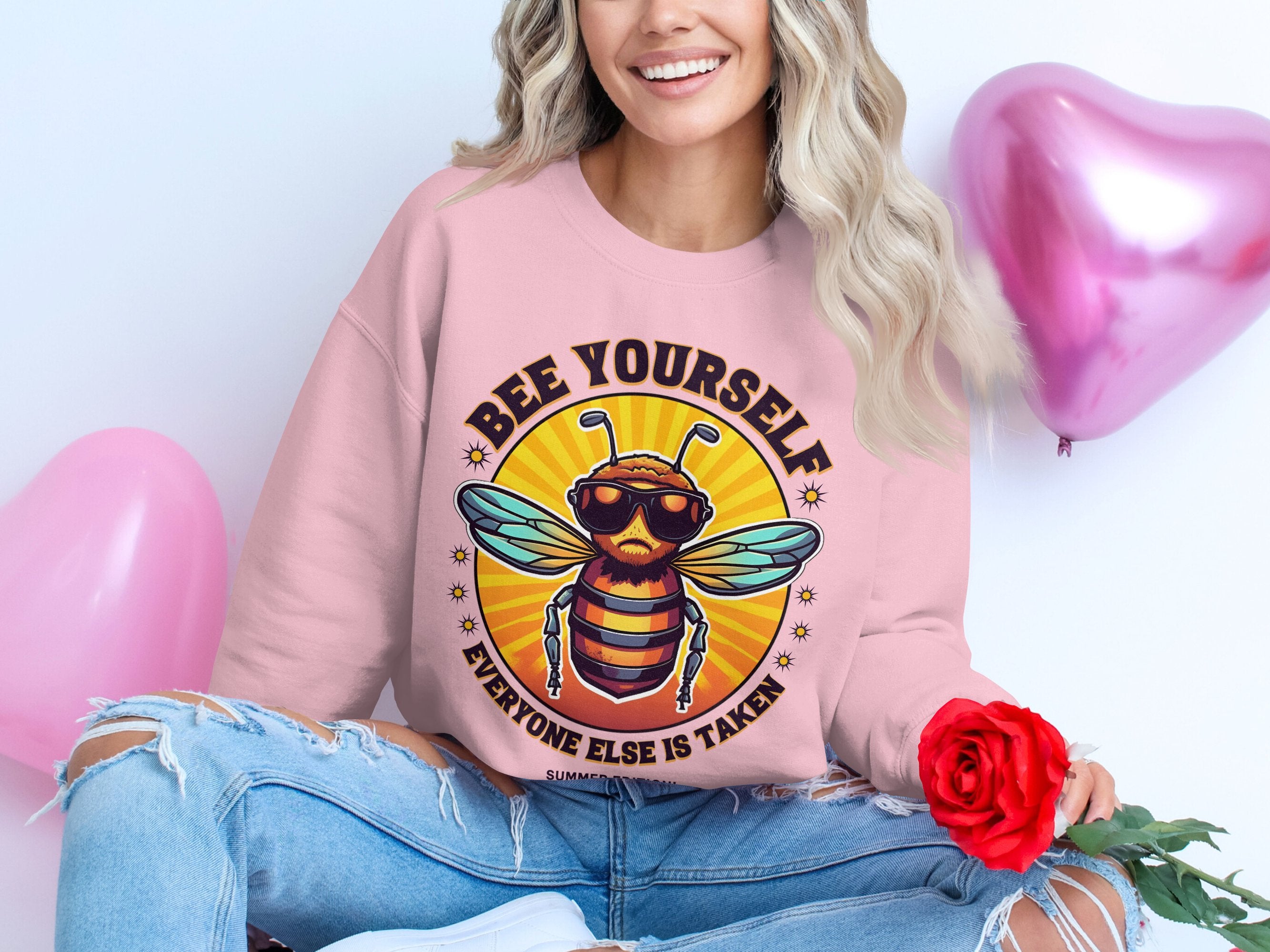 A person holds a red rose while wearing a Garment Graphics pink Bee Yourself, Everyone Else is Taken Summer Edition sweatshirt. Surrounded by three pink heart-shaped balloons and paired with ripped blue jeans, they enjoy the comfy fit of unisex sizing.