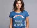 A fishing enthusiast with long dark hair wears a blue Id Rather Have a Bad Day Fishing Than a Good Day at Work T-shirt by Garment Graphics, tailored for women. The shirt features a colorful design and she stands against a neutral gray background.