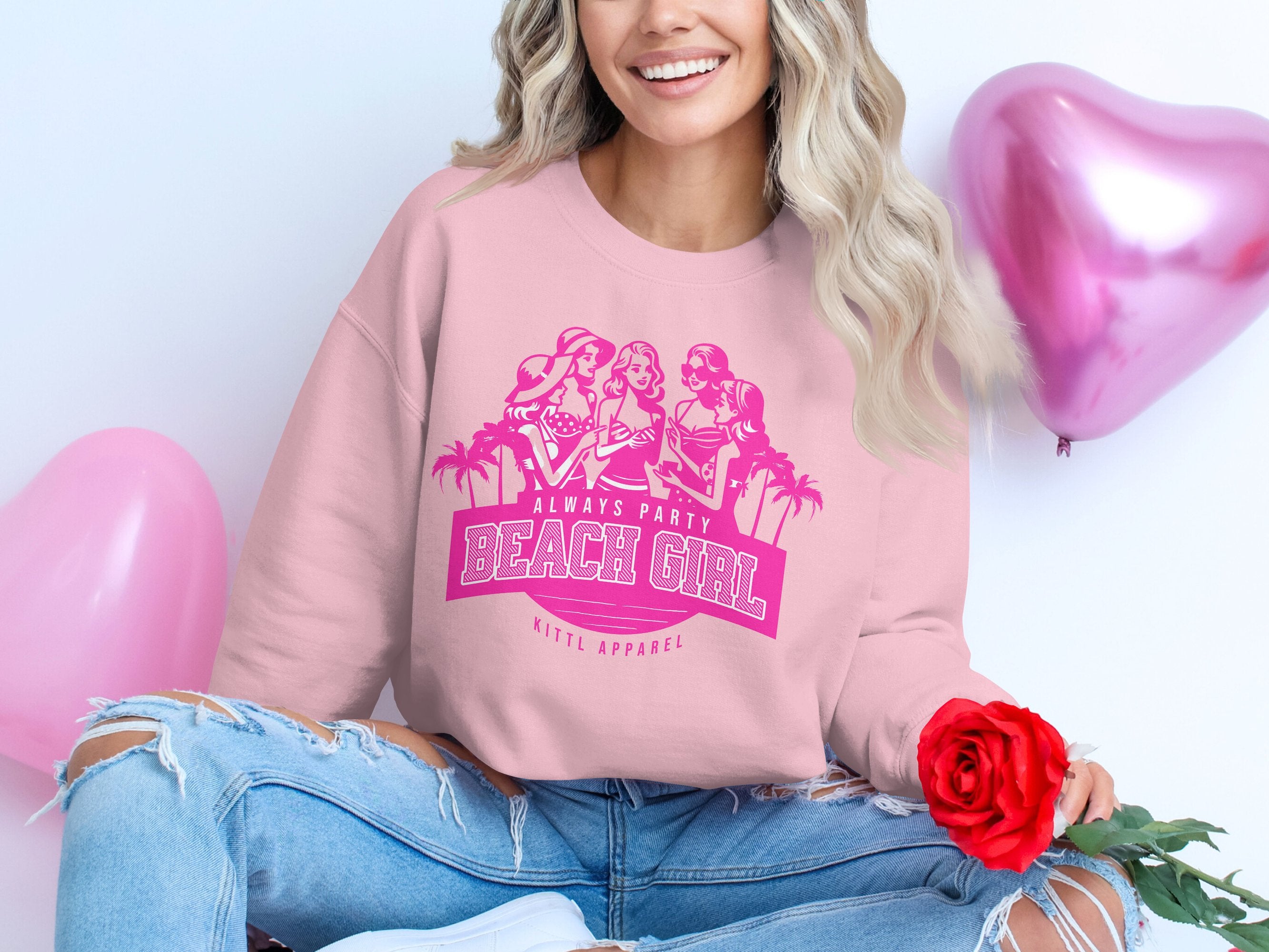 A woman sits cross-legged on the floor, dressed in a pink Garment Graphics sweatshirt featuring cartoons and Always Party Beach Girl text. She holds a red rose by heart-shaped pink balloons, her long wavy hair flowing over ripped jeans.