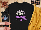 A vintage style black sweatshirt by Garment Graphics boasts a Soccer Mum text and soccer ball graphic. Its showcased with a striking yellow blazer, pink shoes, sunglasses, makeup brushes, vibrant makeup palette, lipsticks, and a small blue pouch on a wooden surface.
