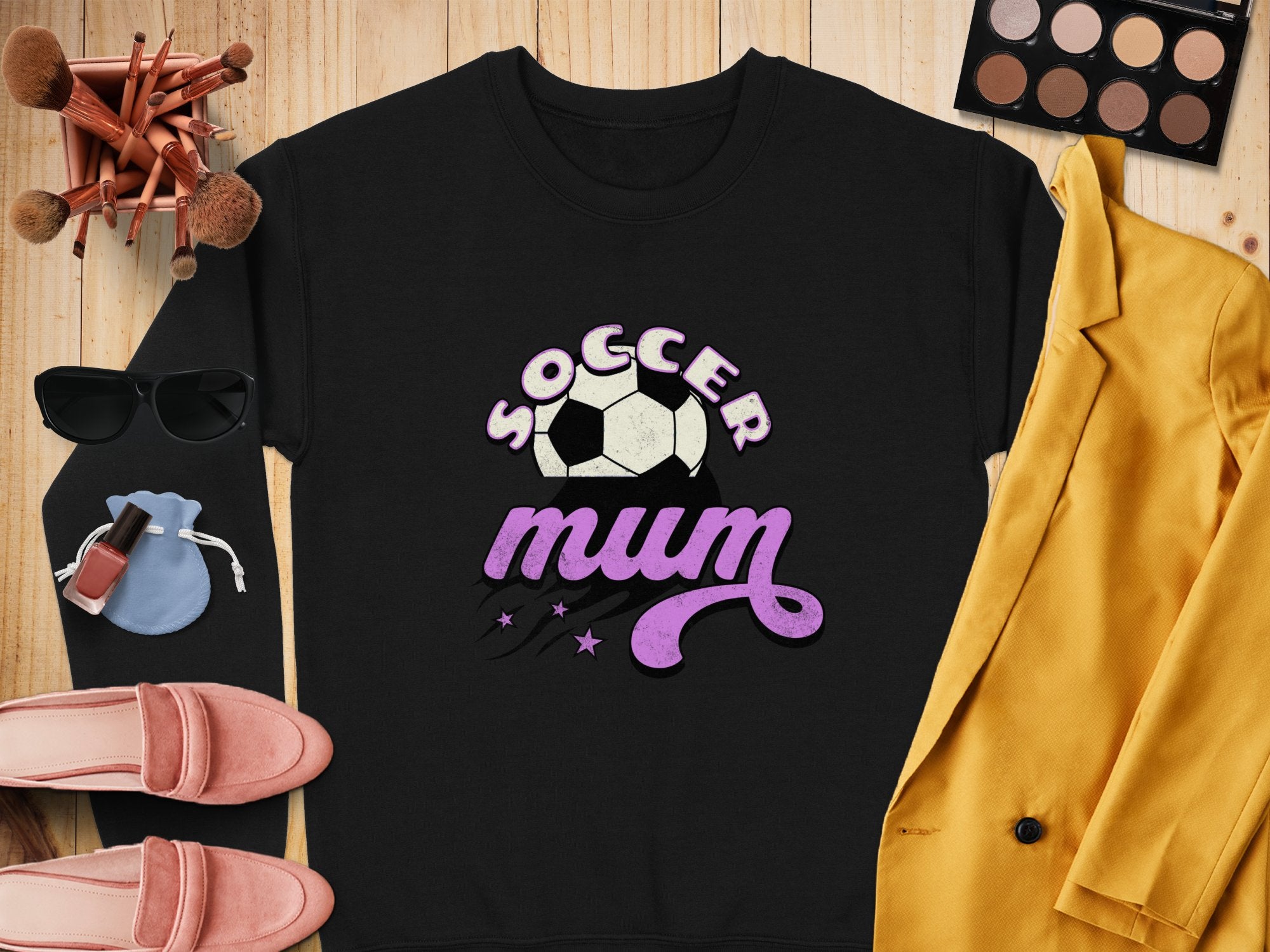 A vintage style black sweatshirt by Garment Graphics boasts a Soccer Mum text and soccer ball graphic. Its showcased with a striking yellow blazer, pink shoes, sunglasses, makeup brushes, vibrant makeup palette, lipsticks, and a small blue pouch on a wooden surface.