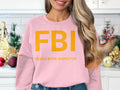 A person in a Garment Graphics sweatshirt featuring FBI Female Boob Inspector in bold yellow relaxes in a festively decorated kitchen with ornaments and mini Christmas trees.