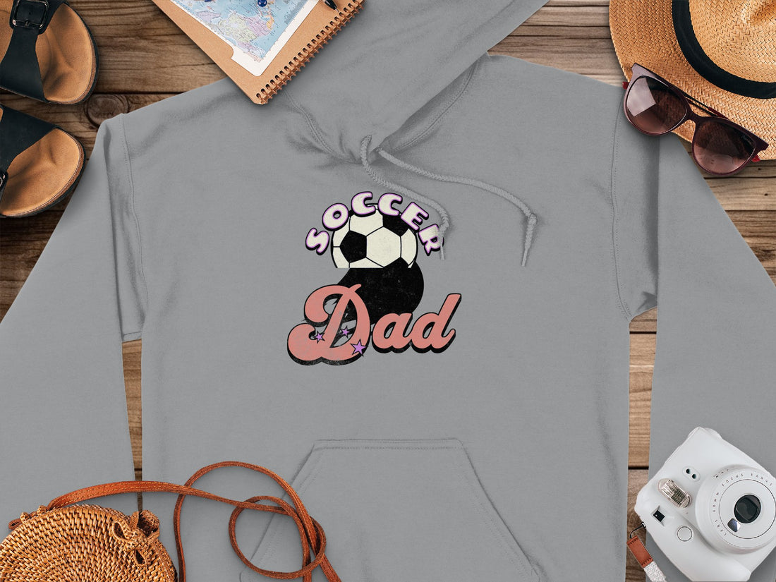 A stylish white hoodie from Garment Graphics features a Soccer Dad print with a soccer ball, displayed on wood beside a hat, sunglasses, notepad, sandals, and camera. Ideal for game days or casual outings.