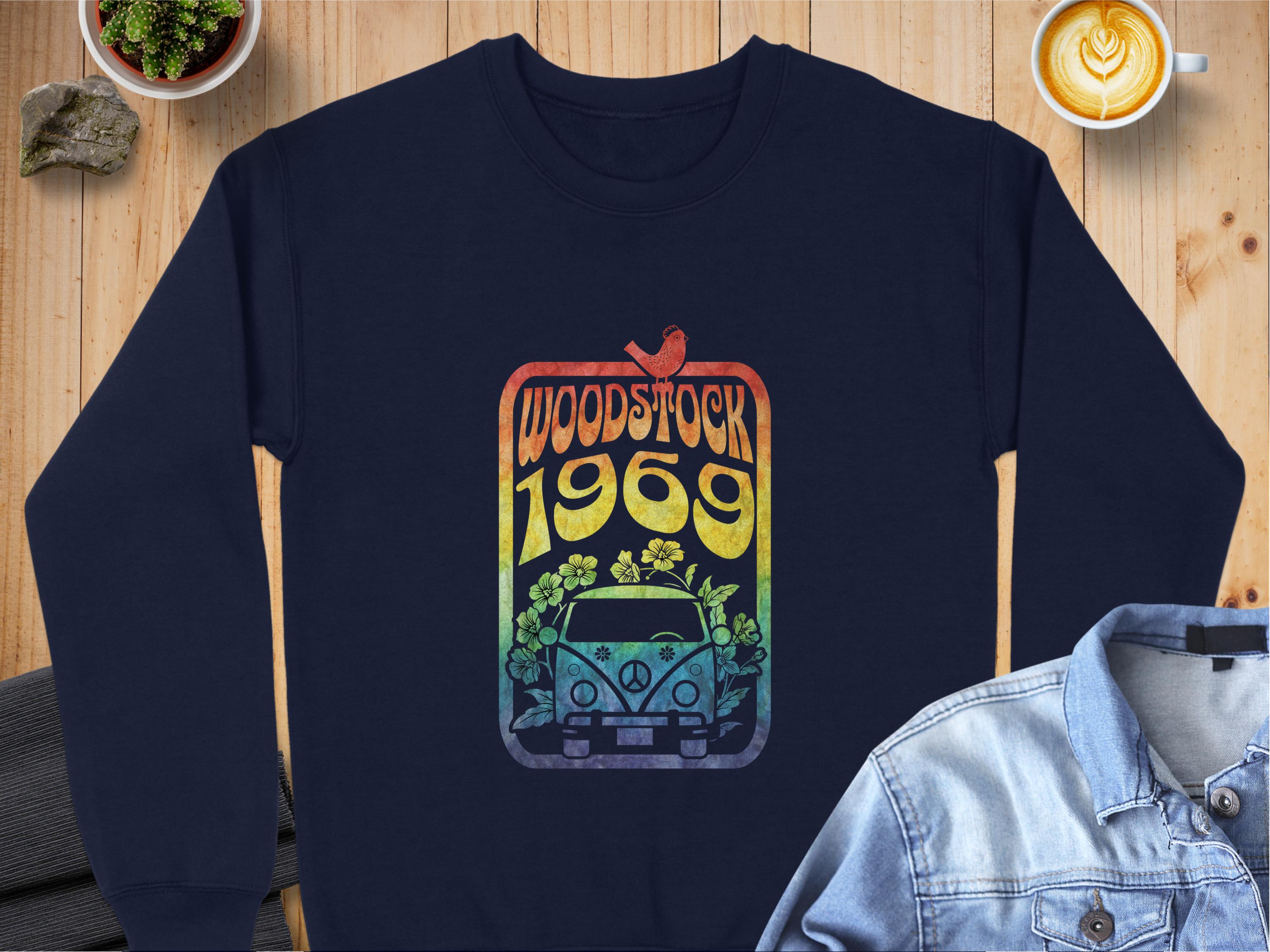 Garment Graphics navy blue sweatshirt has a retro vibe with a vintage-style van, flowers, and bird design. Woodstock 1969 is in rainbow colors. Photographed beside a latte and denim jacket, it captures the legendary Woodstock festivals essence.