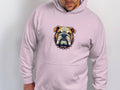 Dressed in a Garment Graphics light pink hoodie featuring a spiked collar bulldog design, the individual gazes downward with one hand casually in their pocket against a plain white background.