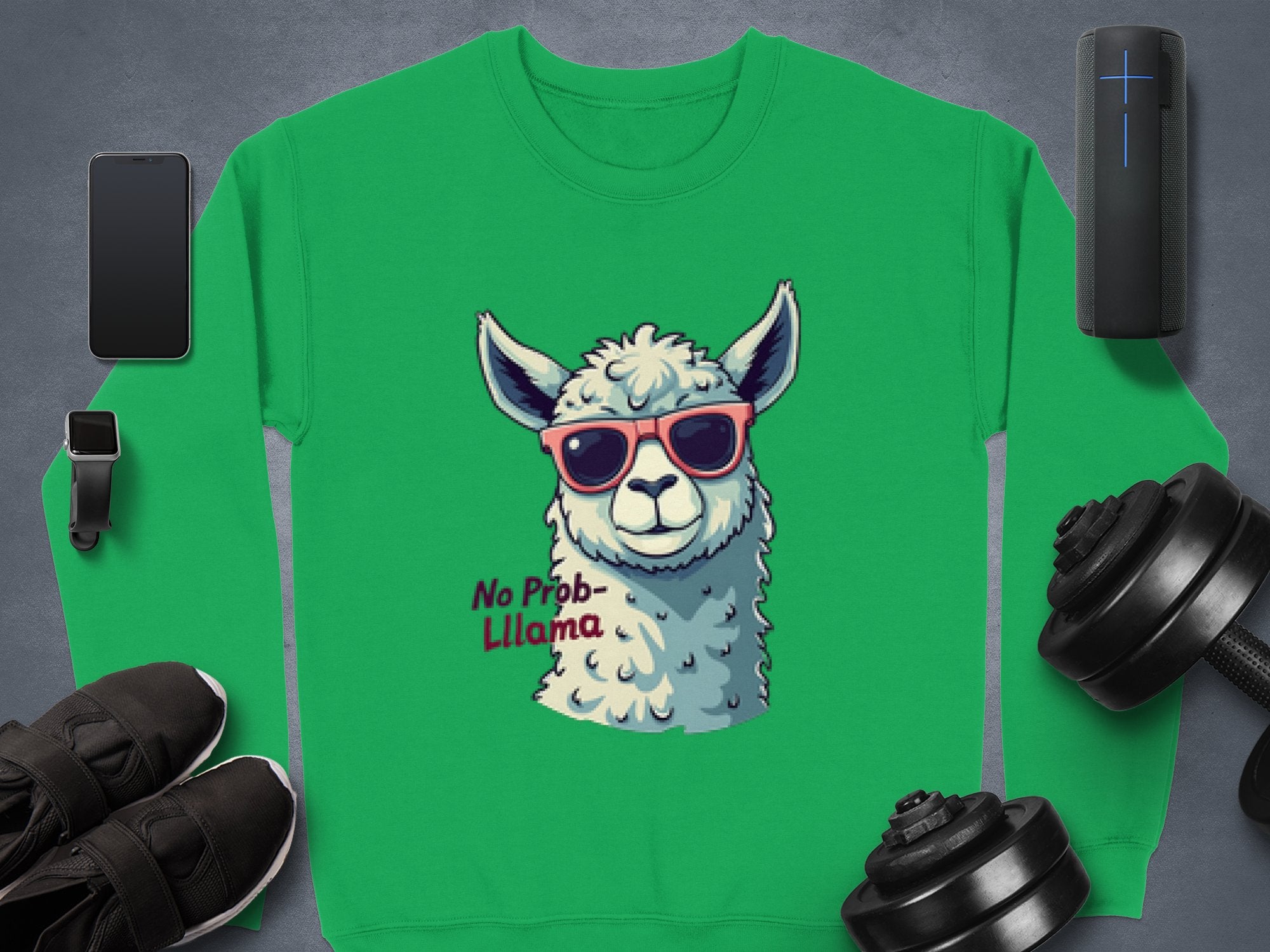 The Garment Graphics unisex sweatshirt features a green llama in red sunglasses with the text No Prob-Llama. Displayed amid a smartwatch, smartphone, wireless speaker, dumbbells, athletic shoes, and a small plant on a gray background.