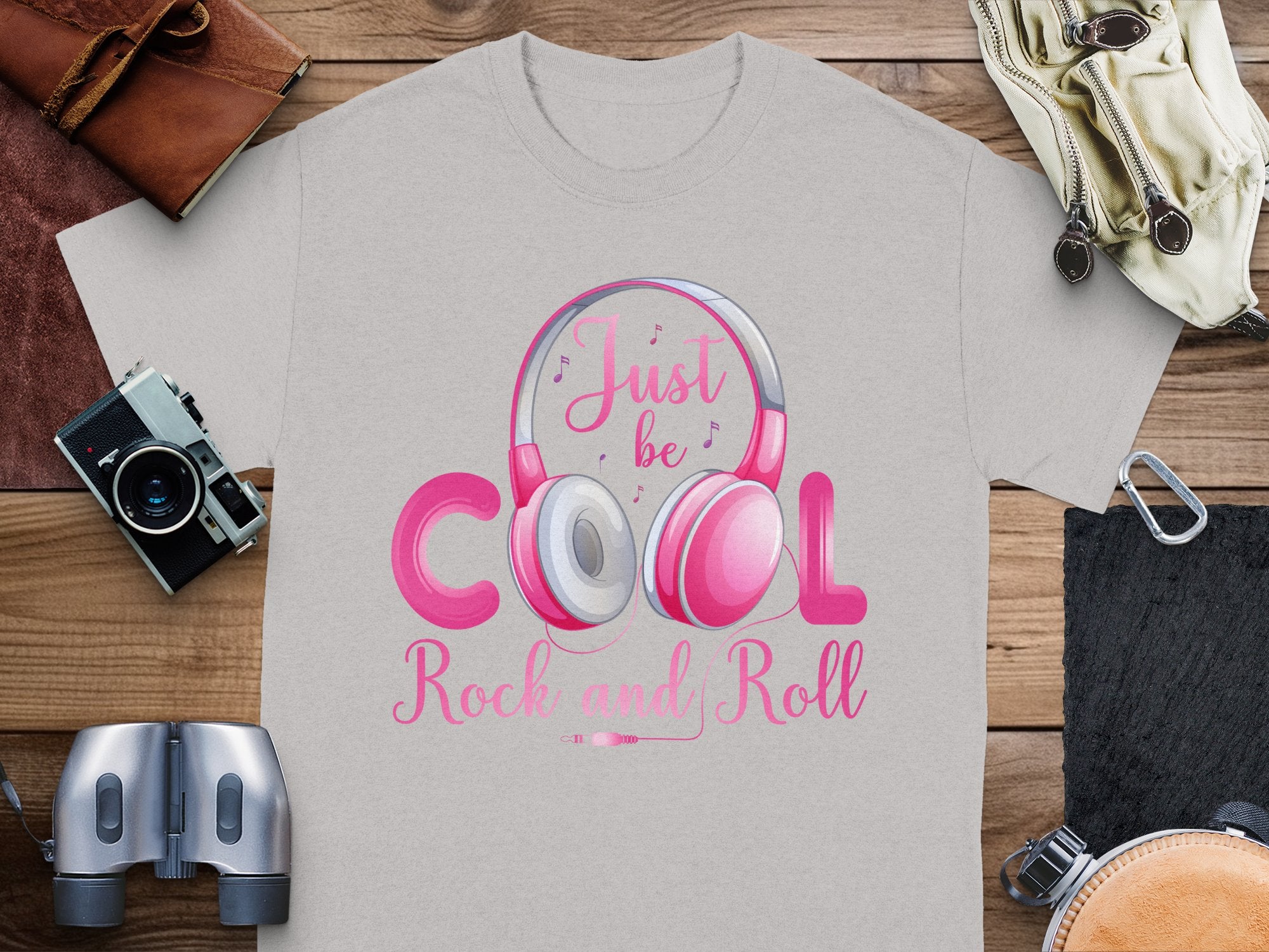 Garment Graphics gray T-shirt showcases headphones and Just be Cool Rock and Roll text. Ideal for music lovers, it pairs well with a camera, binoculars, gloves, leather case, and rope on wood. Perfect for rock and roll enthusiasts.