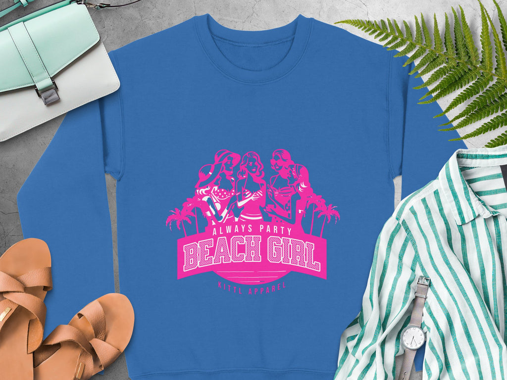 Blue sweatshirt with pink design featuring three women, palm trees, and Always Party Beach text by Garment Graphics. Nearby items include a green-striped shirt, brown sandals, a watch, and a light blue handbag on a concrete surface.