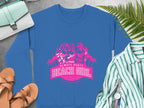 Blue sweatshirt with pink design featuring three women, palm trees, and Always Party Beach text by Garment Graphics. Nearby items include a green-striped shirt, brown sandals, a watch, and a light blue handbag on a concrete surface.