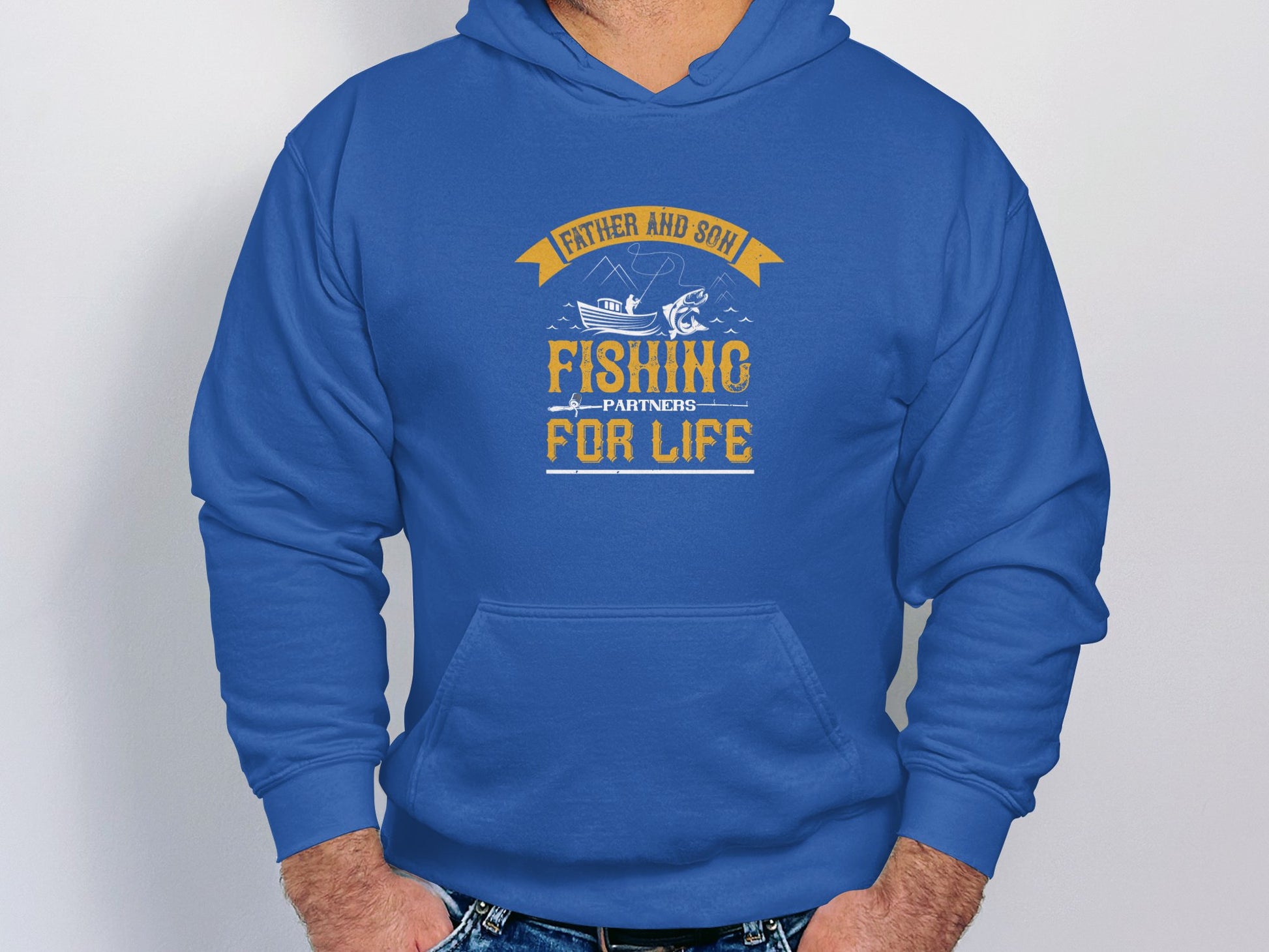 A man models Garment Graphics blue fishing hoodie with a Father and Son, Fishing Partners for Life design featuring a fish and rod, appealing to fishing enthusiasts. His hands rest casually in his jeans pockets.