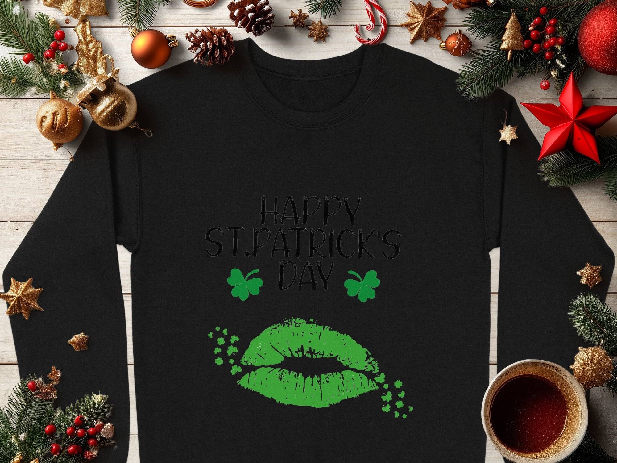The Garment Graphics black sweatshirt showcases a green lips print with Happy St. Patricks Day text, clover kisses, and shamrocks. Set on wood with festive decor, pinecones, and a coffee cup, it exudes a perfect festive spirit.