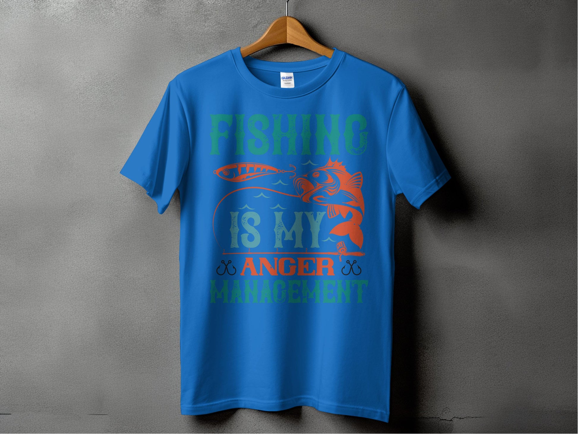 A blue Garment Graphics T-shirt for fishing enthusiasts hangs on a wooden hanger against a gray backdrop. It features bold text and graphics stating Fishing is my anger management alongside a fish and lure design, ideal for those who find peace in fishing.