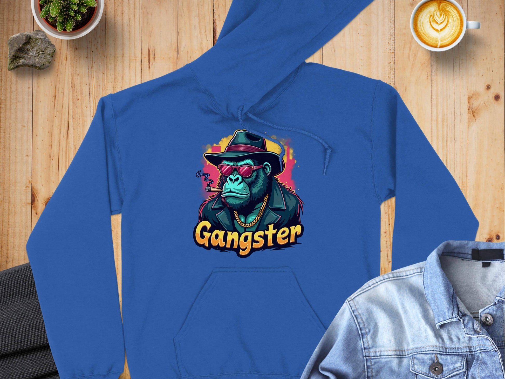 A blue hoodie from Garment Graphics showcases a smokey style with a chimp wearing sunglasses, a hat, and smoking, with Gangster text. Made of medium-heavy fabric, it sits on wood next to a denim jacket, latte, and potted plant.