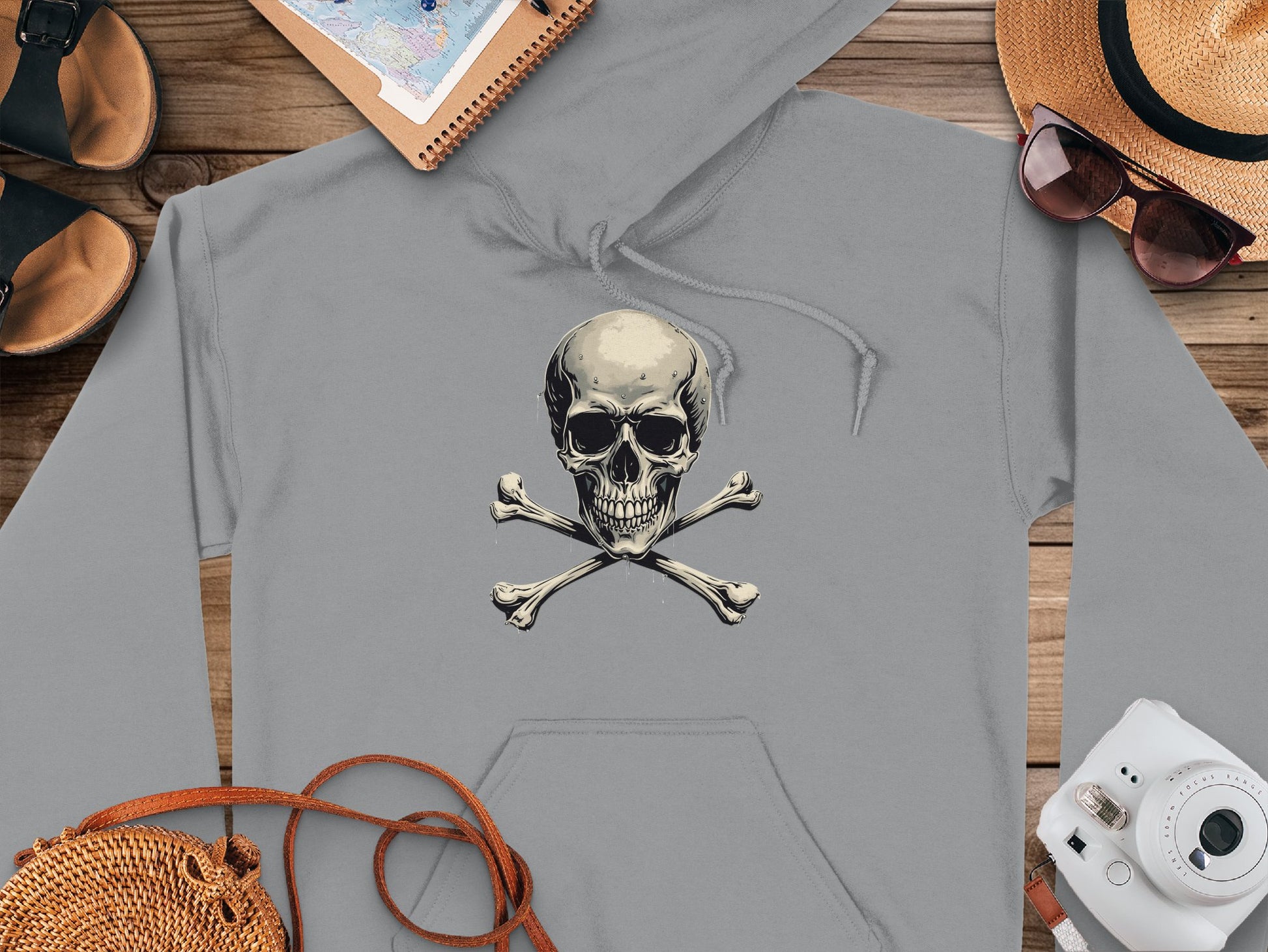 A gray Skull and Crossbones Hoodie from Garment Graphics rests on a wooden surface, exuding chill streetwear vibes. Nearby, a straw hat paired with sunglasses, sandals, a notebook, and a small white camera complete the laid-back look.