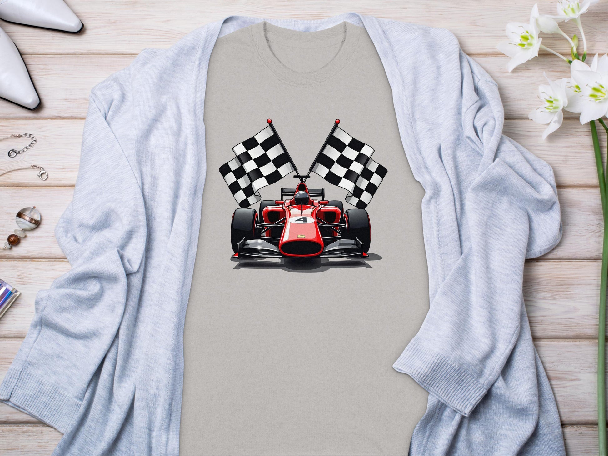 A Garment Graphics gray T-shirt displaying a red racing car and two checkered flags captures the motorsport thrill. Its laid on wood and paired with a light blue cardigan adorned with white and pink flowers on the right.