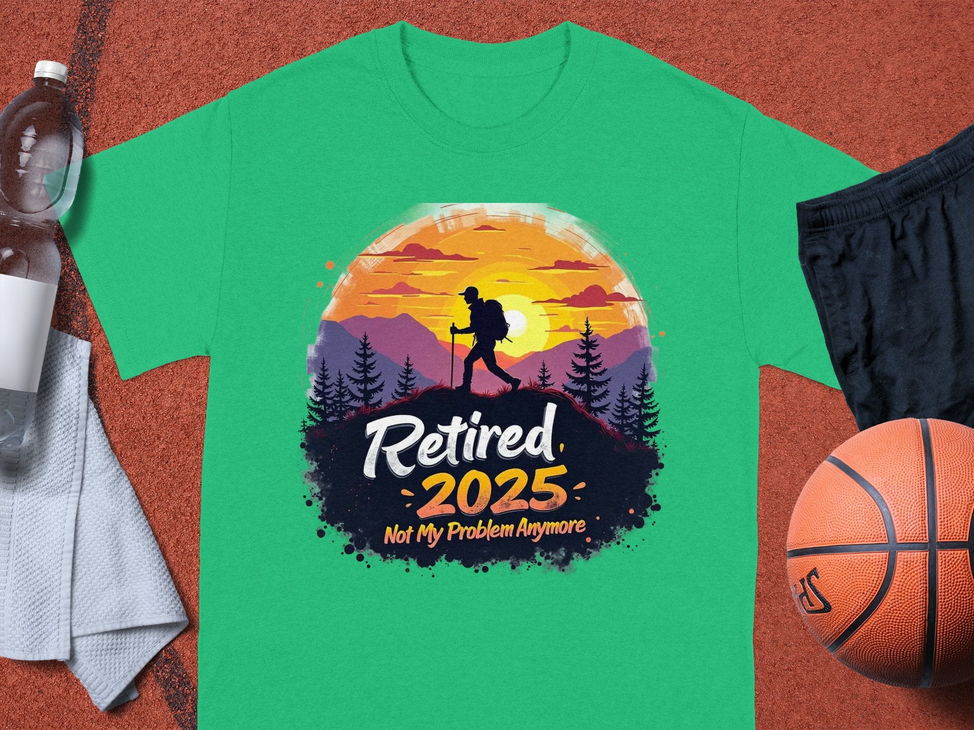 Garment Graphics Green Sunset T-shirt showcases a hiking silhouette against a sunset backdrop with Retired 2025, Not My Problem Anymore text. Laid on an orange surface with a water bottle, towel, and basketball—ideal for celebrating retirement in style.
