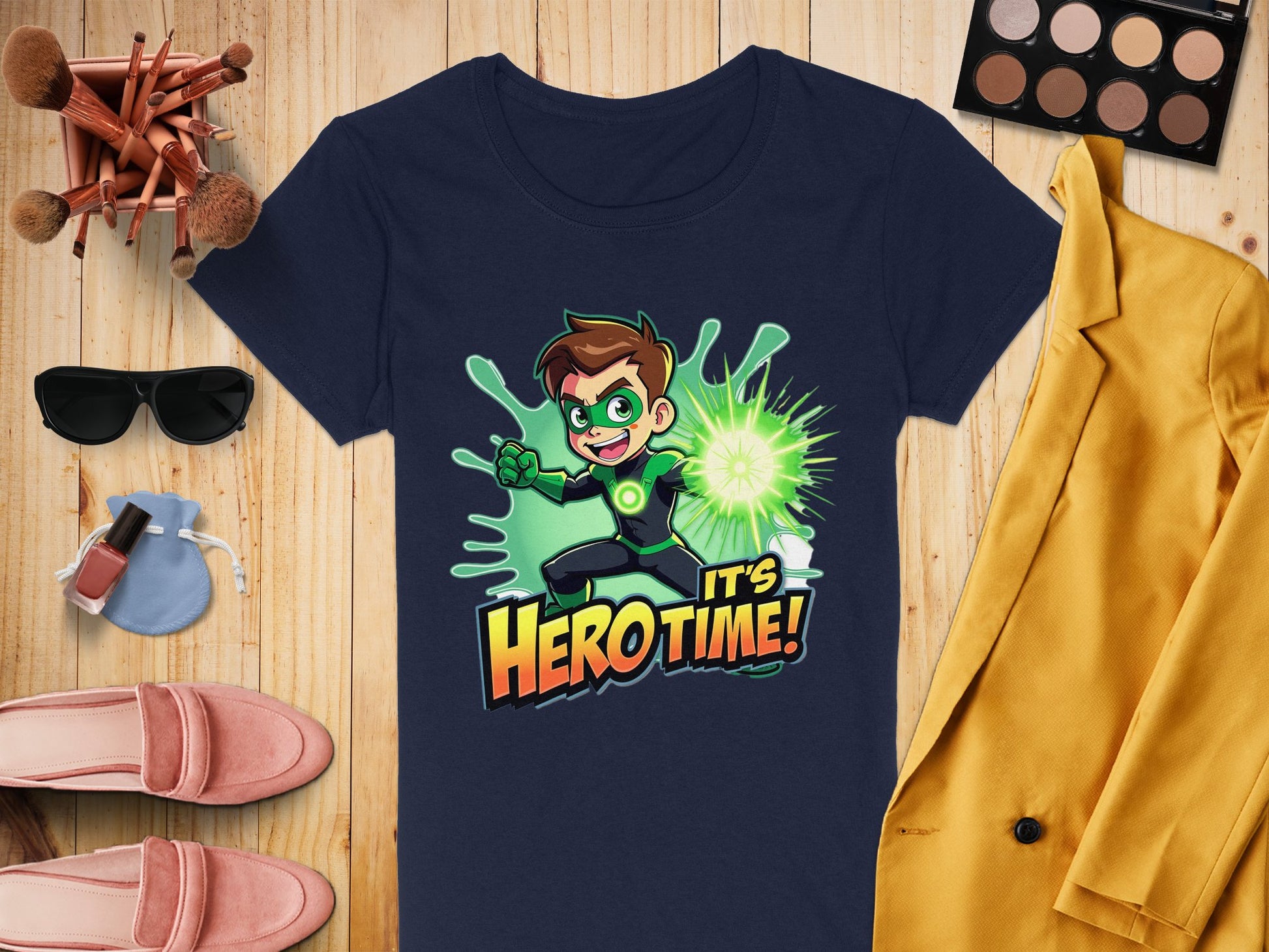 A Garment Graphics navy blue T-shirt with a green superhero and Its Hero Time! text is displayed on wood. Accompanying this are a yellow jacket, eyeshadow palette, makeup brushes, sunglasses, pink loafers, and nail polish.