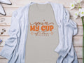 A beige 100% cotton graphic tee by Garment Graphics, featuring Youre My Cup of Tea in orange, is showcased on a light wood surface. Its framed by a cozy light gray cardigan, paired with white high-heeled shoes, delicate jewelry, and fresh white flowers.