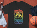 A black Garment Graphics T-shirt with a retro Woodstock 1969 design featuring a van, flowers, and a bird is displayed on a textured surface. Nearby are essentials: a white towel, water bottle, black shorts, and basketball—a perfect ensemble for any music lovers collection.