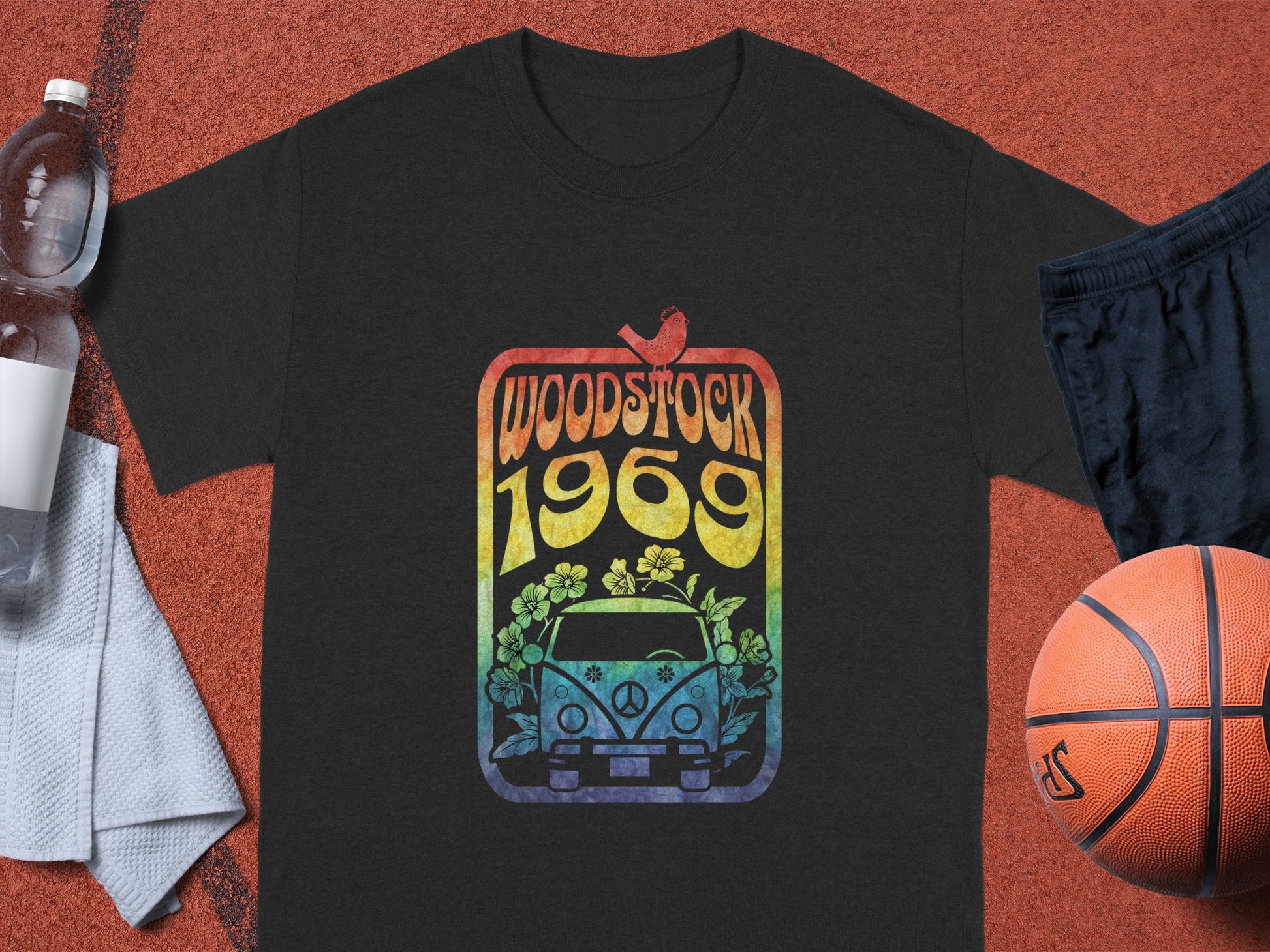 A black Garment Graphics T-shirt with a retro Woodstock 1969 design featuring a van, flowers, and a bird is displayed on a textured surface. Nearby are essentials: a white towel, water bottle, black shorts, and basketball—a perfect ensemble for any music lovers collection.