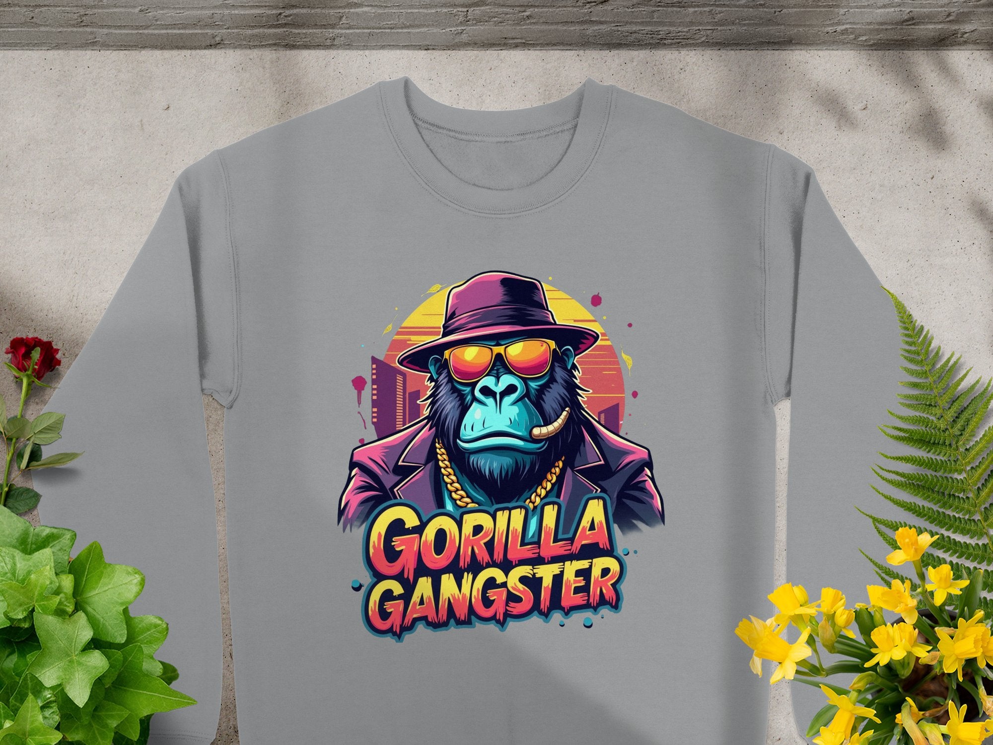 The Garment Graphics urban style gray sweatshirt showcases a gorilla in a hat, sunglasses, gold chain, and cigar, with Gorilla Gangster boldly printed below. Ideal for an edgy look when paired with potted plants on a concrete surface.