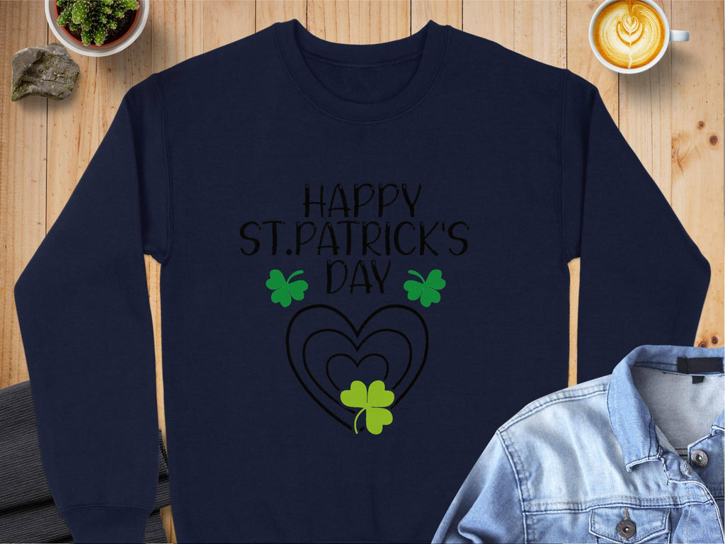 A navy festive sweatshirt from Garment Graphics, featuring Happy St. Patricks Day in black above heart designs and green clovers, is on a wooden table next to a denim shirt, latte, potted plant, and small rock.