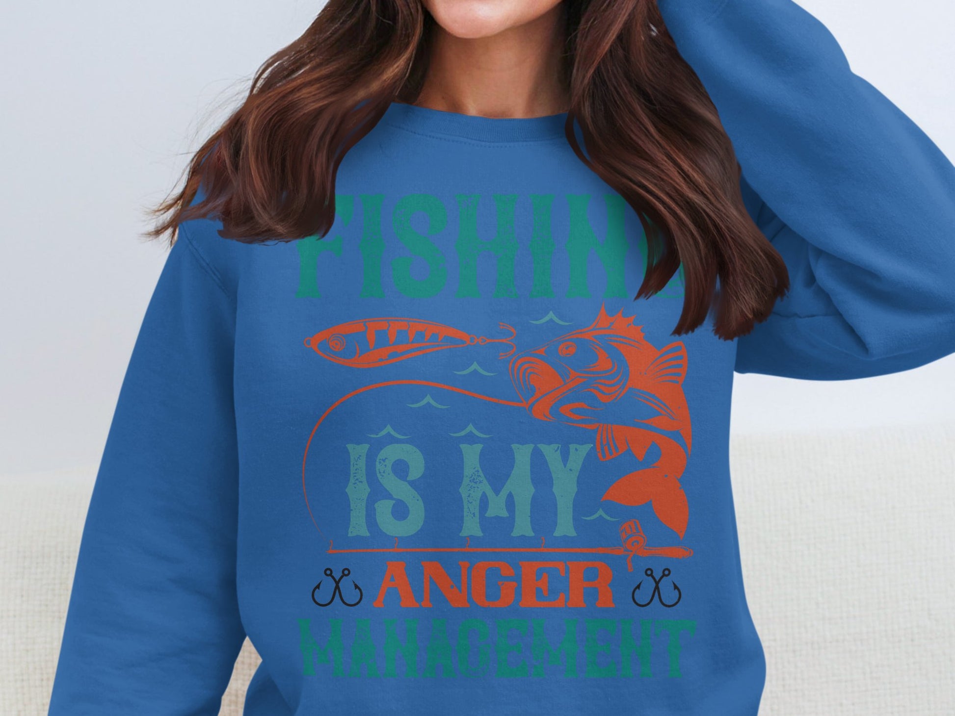 A person sports a blue Garment Graphics sweatshirt with a distinctive fish and lure design. Its ideal for fishing lovers, with Fishing Is My Anger Management emblazoned in bold green, orange, and red letters.