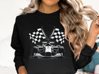 A person with long wavy hair sports a standout design by Garment Graphics—a black sweatshirt featuring a graphic of a Formula 1 car in front of crossed checkered flags, set against a plain light-colored background.