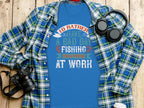 A Garment Graphics blue fishing T-shirt with the slogan, Id rather have a bad day fishing than a good day at work, rests on wood and is partially covered by a plaid shirt. Ideal for anglers, it sits beside a camera, headphones, and small plant.