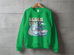 Garment Graphics unisex green sweatshirt showcases a Beach Vibes graphic with a vintage car, surfboard, wave, and palm trees. Ideal for retro road-trip fans, its presented on a wooden hanger against a concrete backdrop.