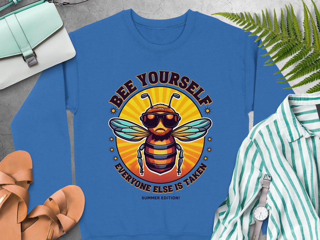 A blue summer edition sweatshirt by Garment Graphics, showcasing a cartoon bee in sunglasses with Bee Yourself, Everyone Else is Taken! text, is displayed with a white and green striped shirt, sandals, a purse, and watch. Its available in unisex sizing for everyone to enjoy.