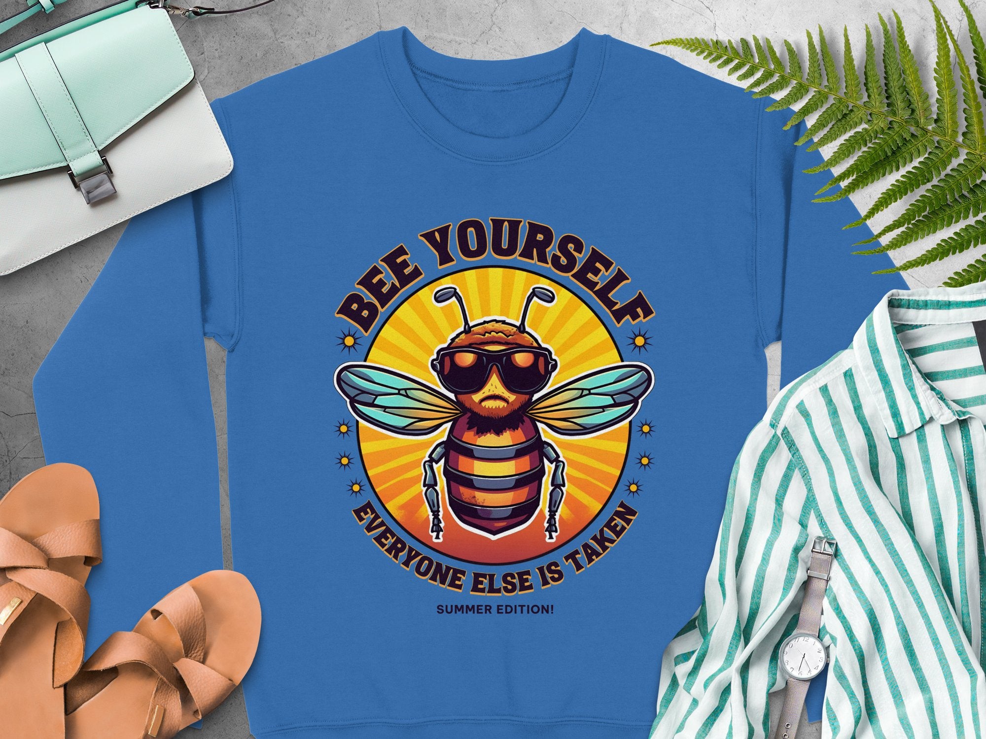 A blue summer edition sweatshirt by Garment Graphics, showcasing a cartoon bee in sunglasses with Bee Yourself, Everyone Else is Taken! text, is displayed with a white and green striped shirt, sandals, a purse, and watch. Its available in unisex sizing for everyone to enjoy.