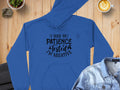 A Garment Graphics blue novelty hoodie with black text, I had my patience tested Im negative, rests on a wooden table with a latte, plant, rocks, and denim jacket nearby. This humorous statement piece adds unique flair to your casual attire.