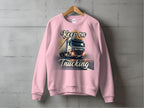 A pink sweatshirt by Garment Graphics hangs on a wooden hanger against a gray wall, featuring a white semi-truck on an open road with Keep on Trucking boldly above and below the image.