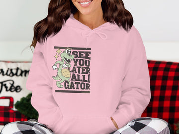 A person wears a classic fit light pink hoodie by Garment Graphics, featuring a gator graphic and the text SEE YOU LATER ALLIGATOR, sitting against a festive checkered background, exuding comfort and style.