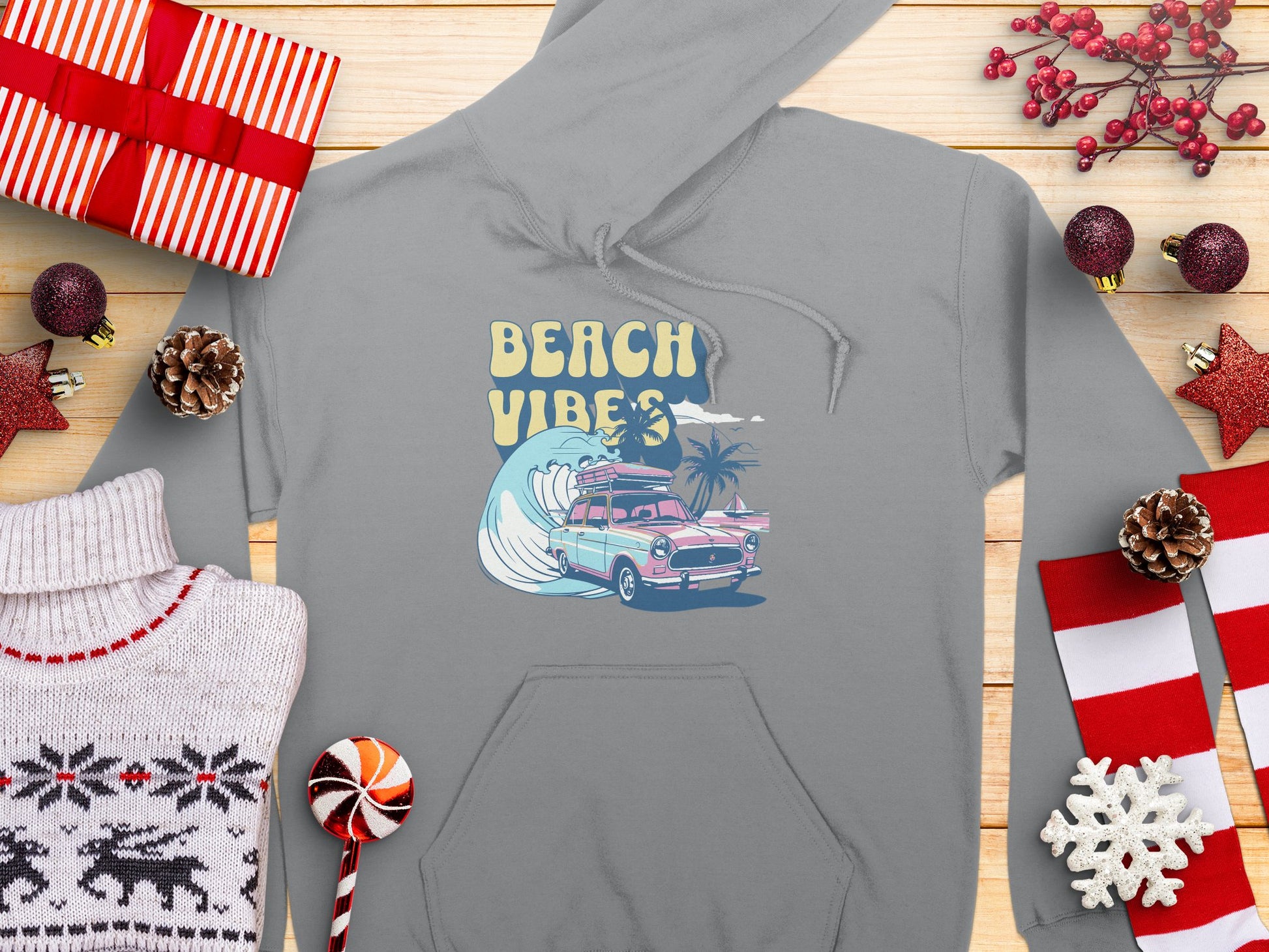 A vibrant gray Garment Graphics hoodie with a Beach Vibes surfboard design, featuring a car and palm trees, is laid on wood. Surrounded by wrapped presents, a reindeer sweater, pine cones, candy, and festive red and white holiday elements, it exudes retro beach style charm.