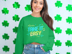A person with long hair stands in front of a wall covered in green shamrocks, wearing a Garment Graphics unisex Take It Easy sweatshirt, featuring musical notes and arrows.