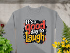 Surrounded by green plants and yellow flowers on a concrete background, the Garment Graphics sweatshirt features medium-heavy gray fabric with Its a good day to laugh in bold, colorful lettering. Its designed for unisex sizing, offering effortless style and comfort.