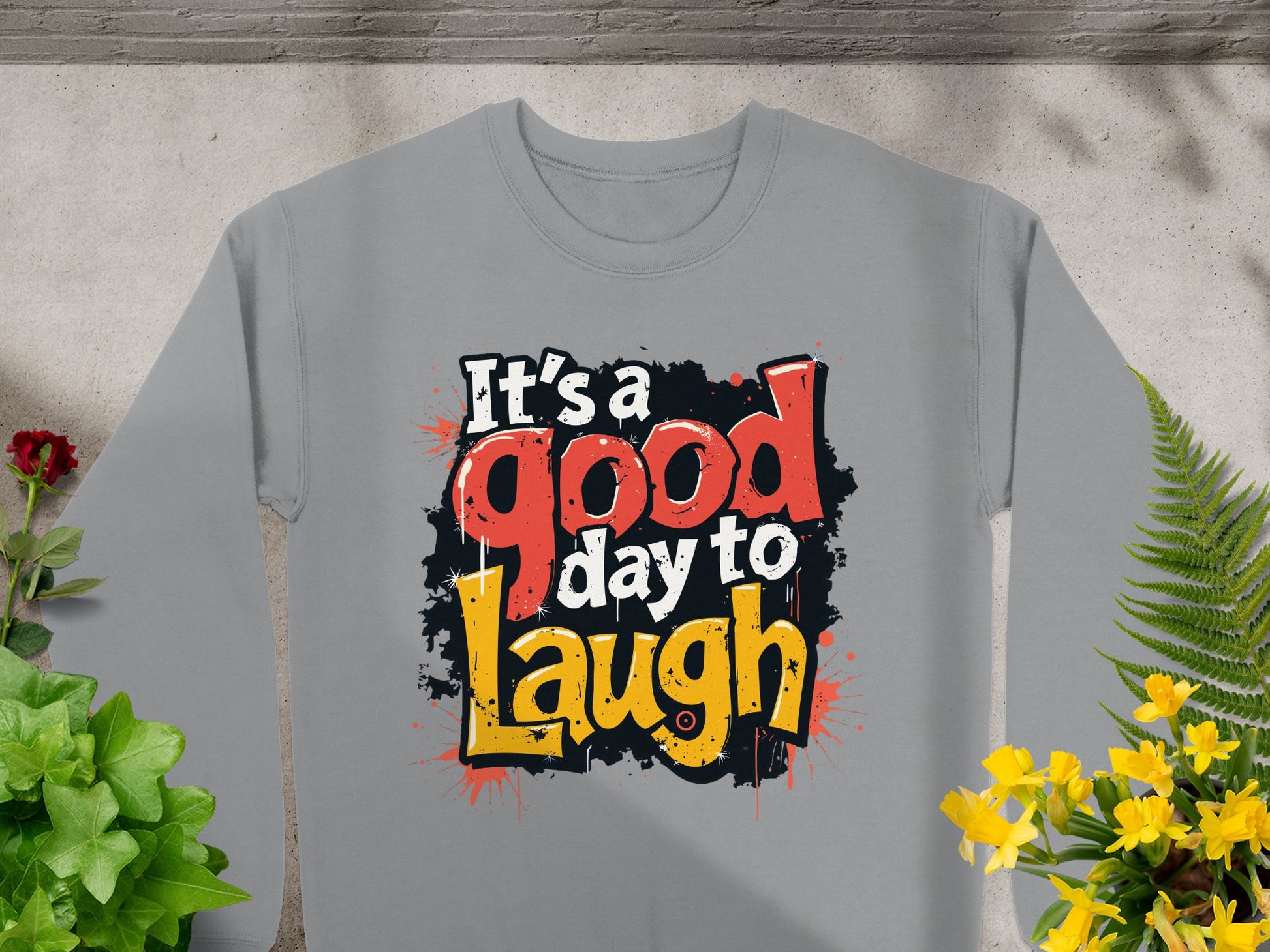 Surrounded by green plants and yellow flowers on a concrete background, the Garment Graphics sweatshirt features medium-heavy gray fabric with Its a good day to laugh in bold, colorful lettering. Its designed for unisex sizing, offering effortless style and comfort.