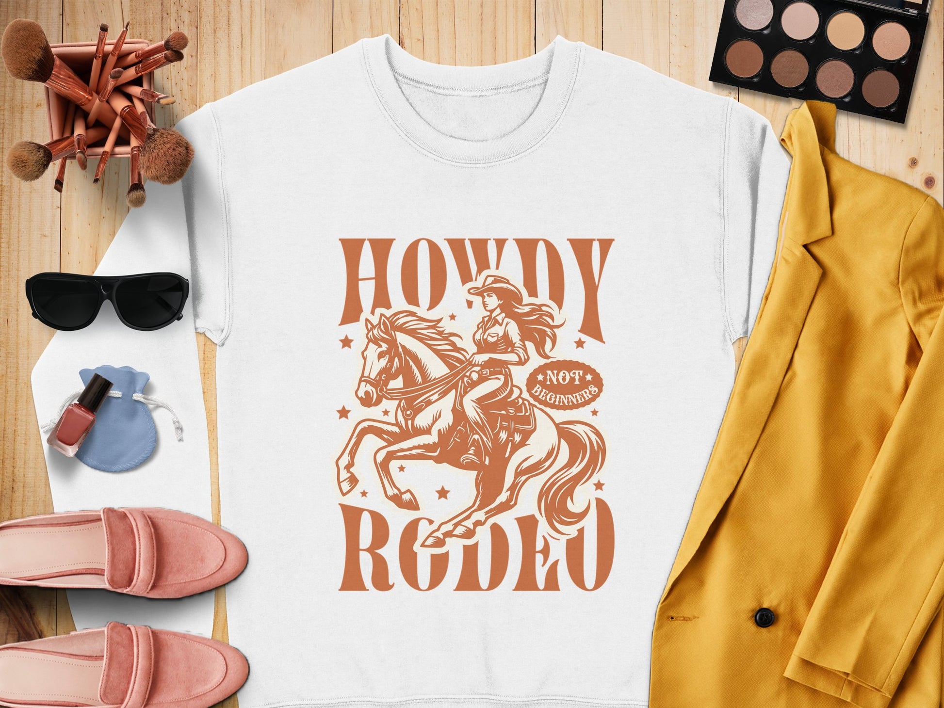 A flat lay image displays Garment Graphics Howdy Rodeo Sweatshirt, featuring a cowgirl on a horse with Not a Beginner. Surrounded by sunglasses, brushes, pink shoes, a mustard yellow blazer, purse, nail polish, and an eye shadow palette, its ideal for rodeo enthusiasts.