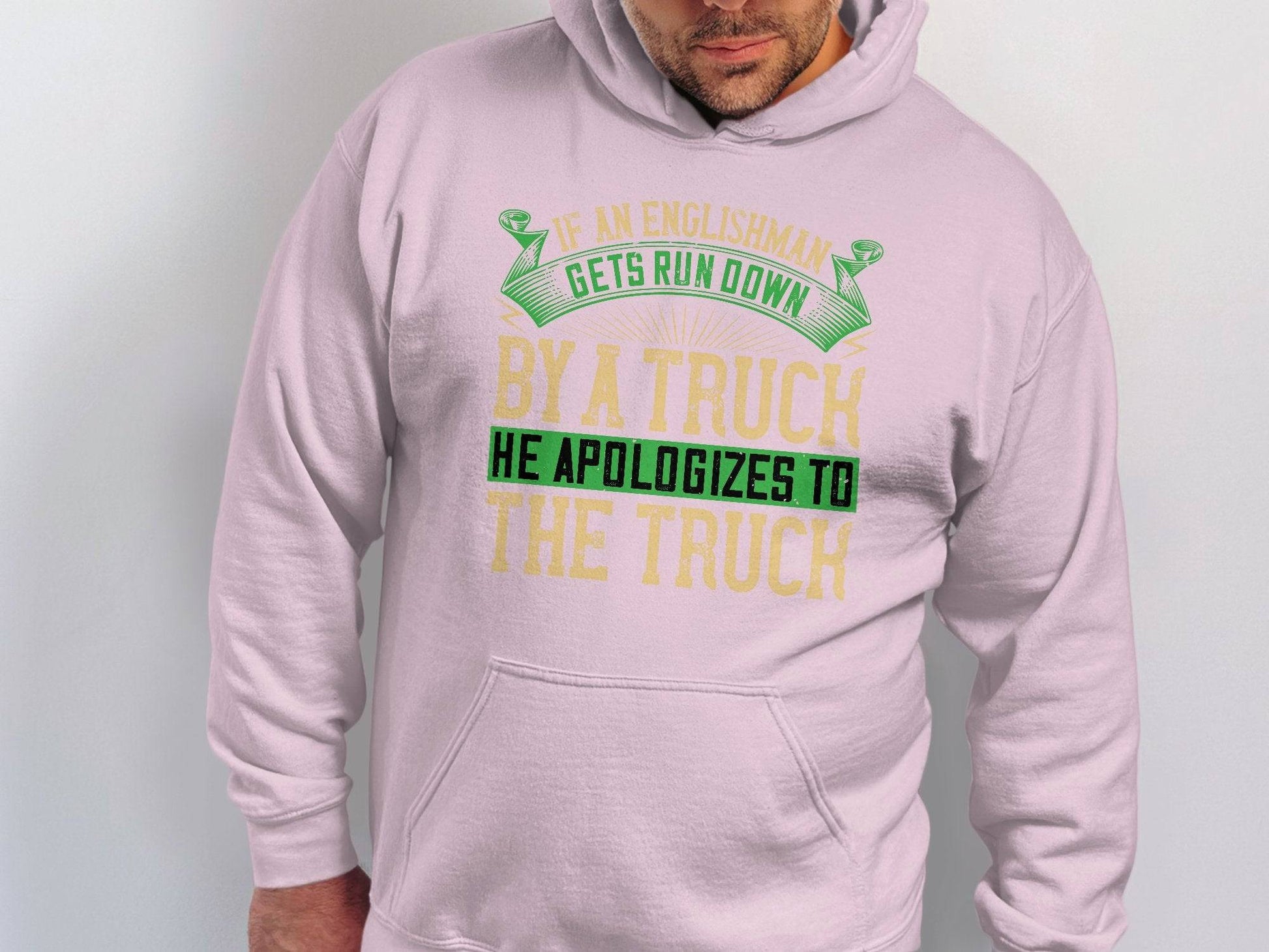 A person wearing a Garment Graphics light pink hoodie sports a green and yellow quote: If an Englishman gets run down by a truck he apologizes to the truck. The humorous text highlights English etiquette as the wearer looks downward.