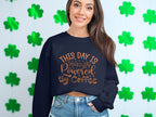A woman wearing a Garment Graphics sweatshirt with This day is powered by coffee in orange stands against green shamrocks, displaying a playful design.