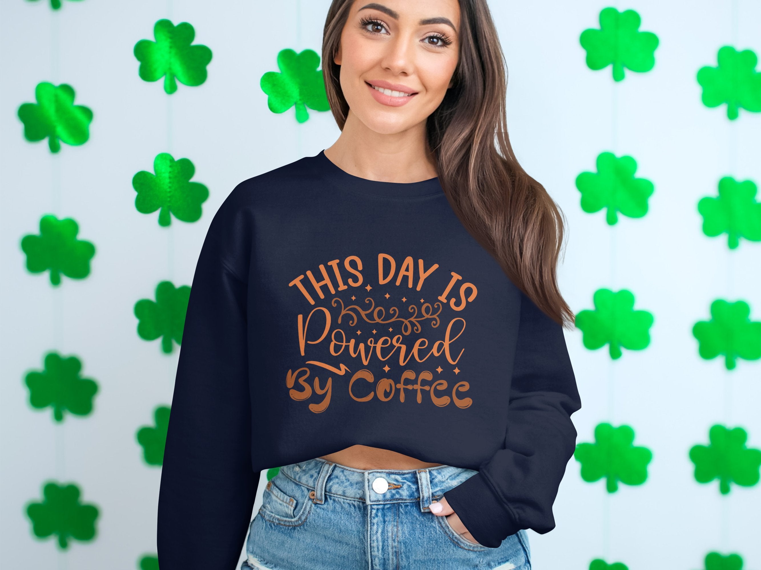 A woman wearing a Garment Graphics sweatshirt with This day is powered by coffee in orange stands against green shamrocks, displaying a playful design.