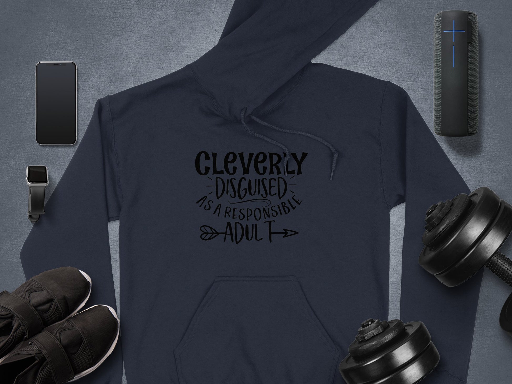 A flat lay image from Garment Graphics features a navy Responsible Adult Hoodies design humorously labeled with Cleverly Disguised as a Responsible Adult, surrounded by a smartphone, wireless speaker, smartwatch, shoes, and dumbbells on a sleek gray background.