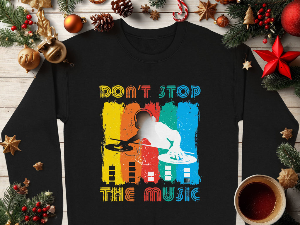 The Garment Graphics black sweater boasts a vivid DJ design with DONT STOP THE MUSIC text, encircled by holiday décor such as ornaments, pine branches, cinnamon sticks, and a coffee cup on wood—ideal for music lovers this season.