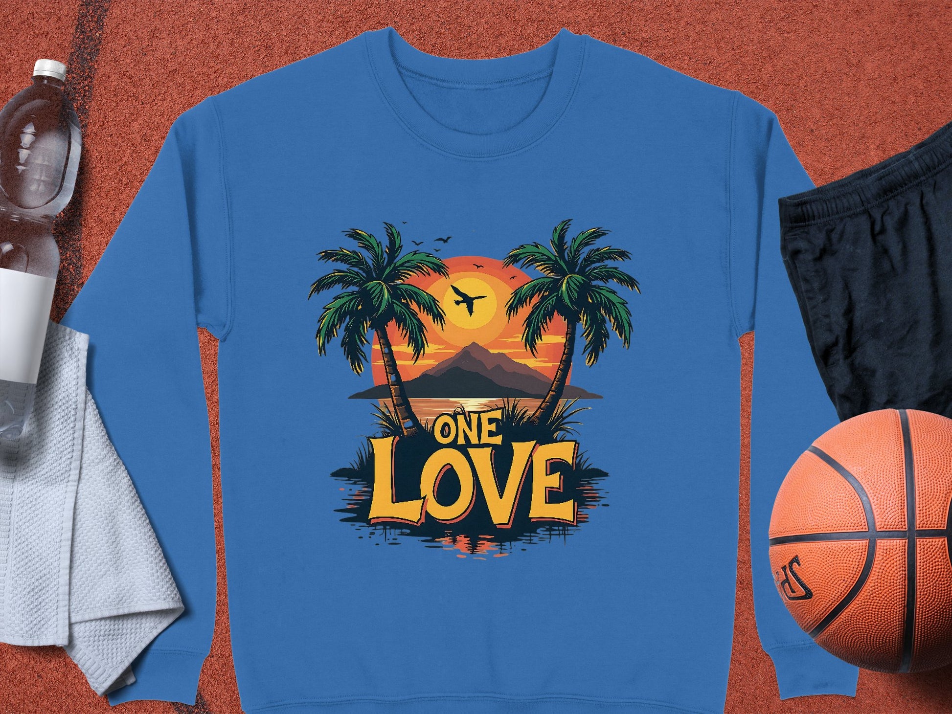 A Garment Graphics blue sweatshirt displays a tropical sunset scene with a mountain, plane, and One Love text. This unisex piece is shown on a textured surface alongside water, a towel, black shorts, and a basketball.